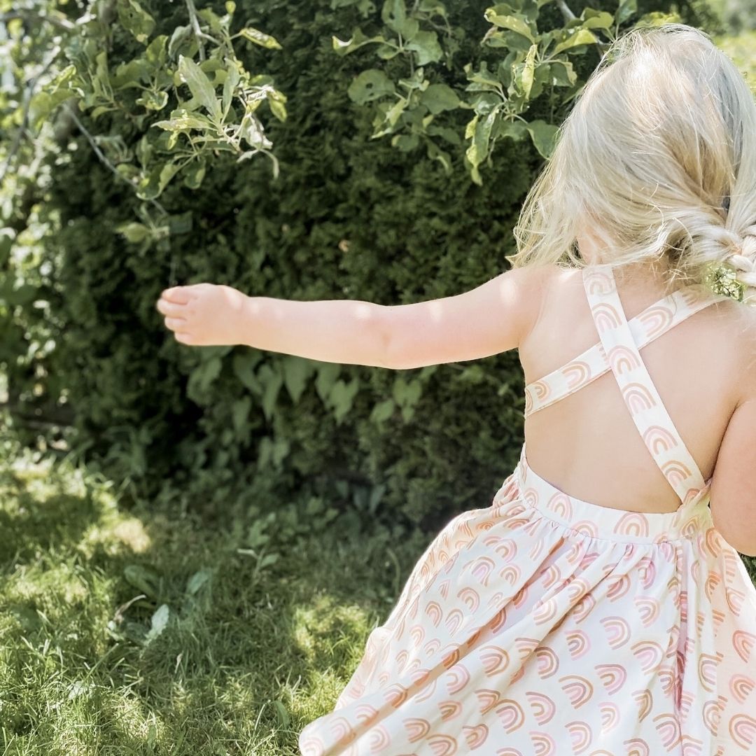 Three year old blonde girl twirling showcasing the back details of criss crossed straps across an open back with a full circle twirl dress. She is wearing the Sofia dress in muted rainbow.