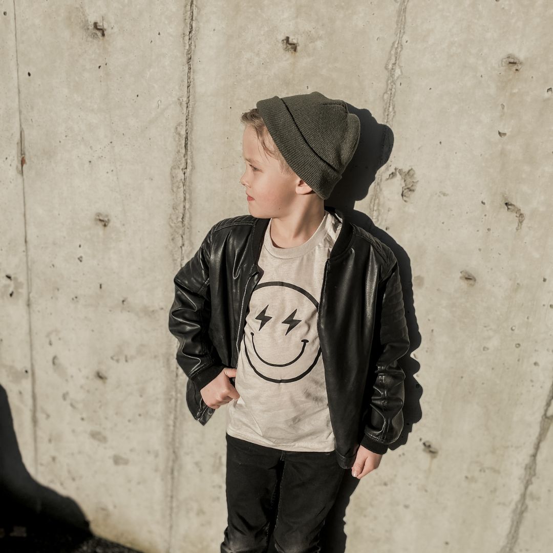 Toddler boy wearing the rad smiley shirt in taupe with black denim jeans, a leather jacket, and a green beanie.