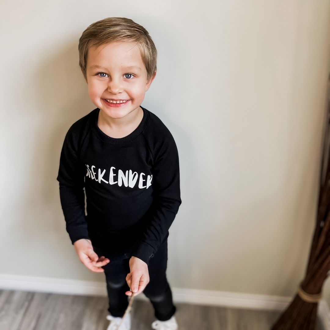 Four year old boy smiling at the camera holding a stick wearing the weekender raglan pullover sweater in black with black jeans.