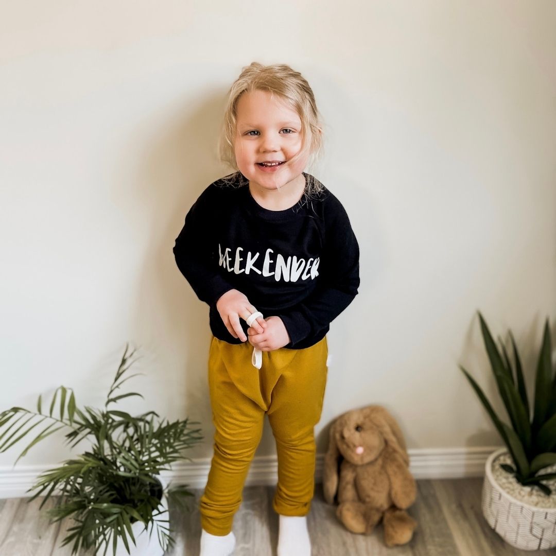 Three year old blonde girl smiling wearing the weekender raglan pullover sweater in black and the harem pants in golden yellow.