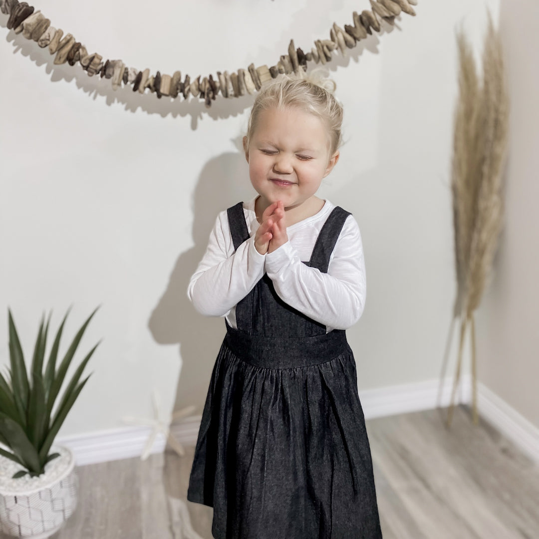 three year old blonde girl wearing the apron dress in black denim over a white long sleeved shirt.