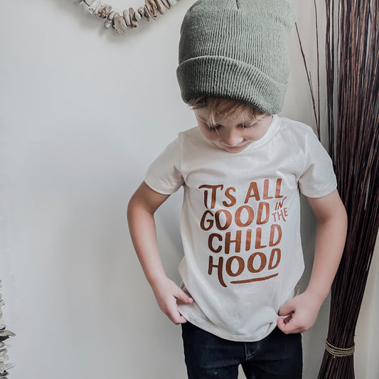 little boy wearing a green toque and a neutral wheat coloured t-shirt with "It's all good in the childhood" written across the front in a camel/caramel coloured print. It is paired with black jeans.