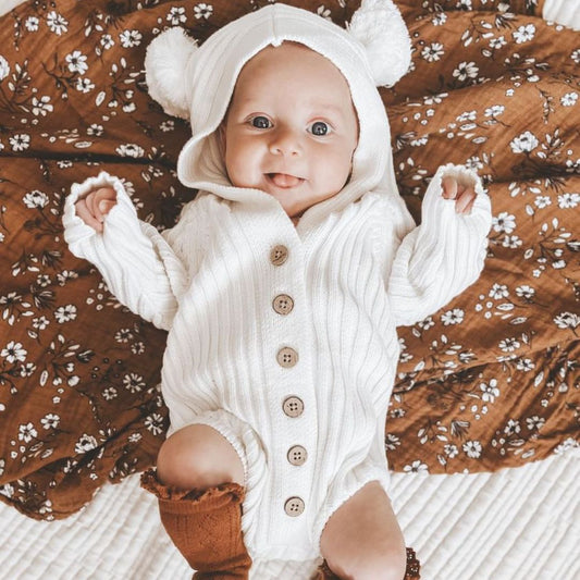 Newborn baby in the hooded knit romper in the color milk. Featuring wood like buttons and adorable pom pom ears.