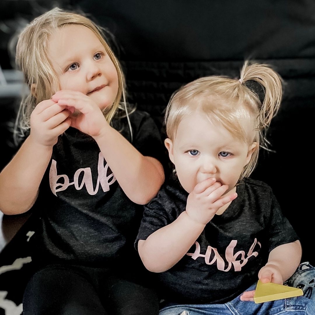 Two sisters sitting on a black chair eating snacks wearing heather black shirts with babe written in pink script on the front.  Edit alt text