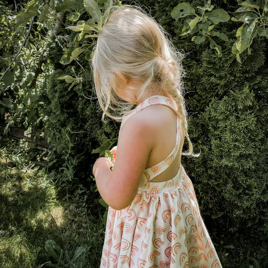 Three year old blonde girl playing with a tree wearing the Sofia dress in muted rainbow.