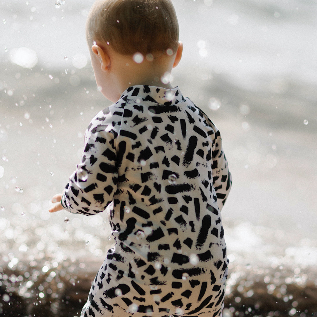 baby approaching the waves with water splashing up. He is wearing a full sleeve rash guard with knee length legs. The sunsuit is white with black paint stripes on it.
