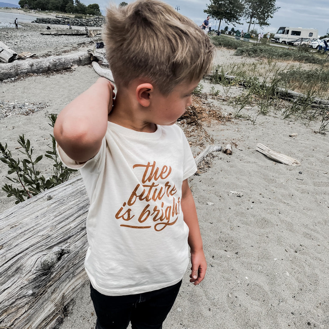 Five year old blond boy standing on a sandy beach wearing the future is bright t-shirt in wheat and black jeans.