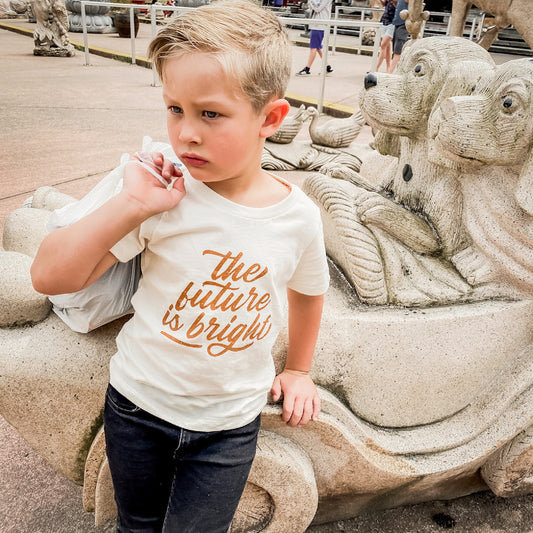 five year old blonde boy leaning against a statue of puppies in a car wearing the future is bright t-shirt in wheat and black jeans.