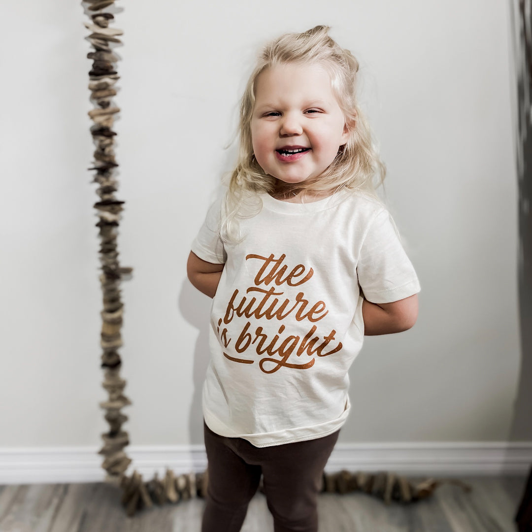 Three year old blonde girl wearing the future is bright t-shirt in wheat with caramel coloured writing and brown leggings.