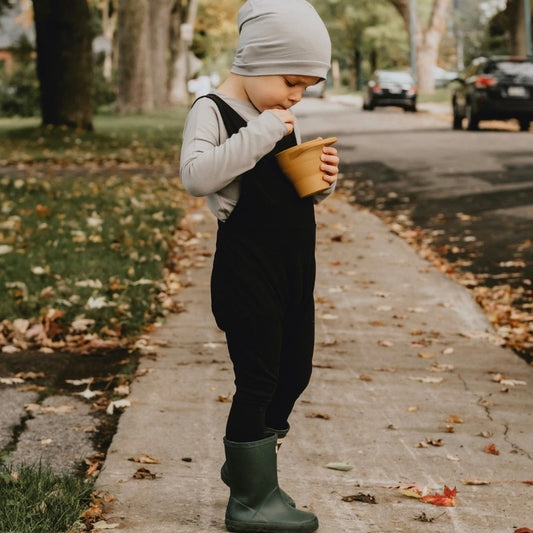little boy eating ice cream out of a terracotta cup wearing the grow with me overalls in black with rain boots, a toque, and the grow with me shirt in taupe.