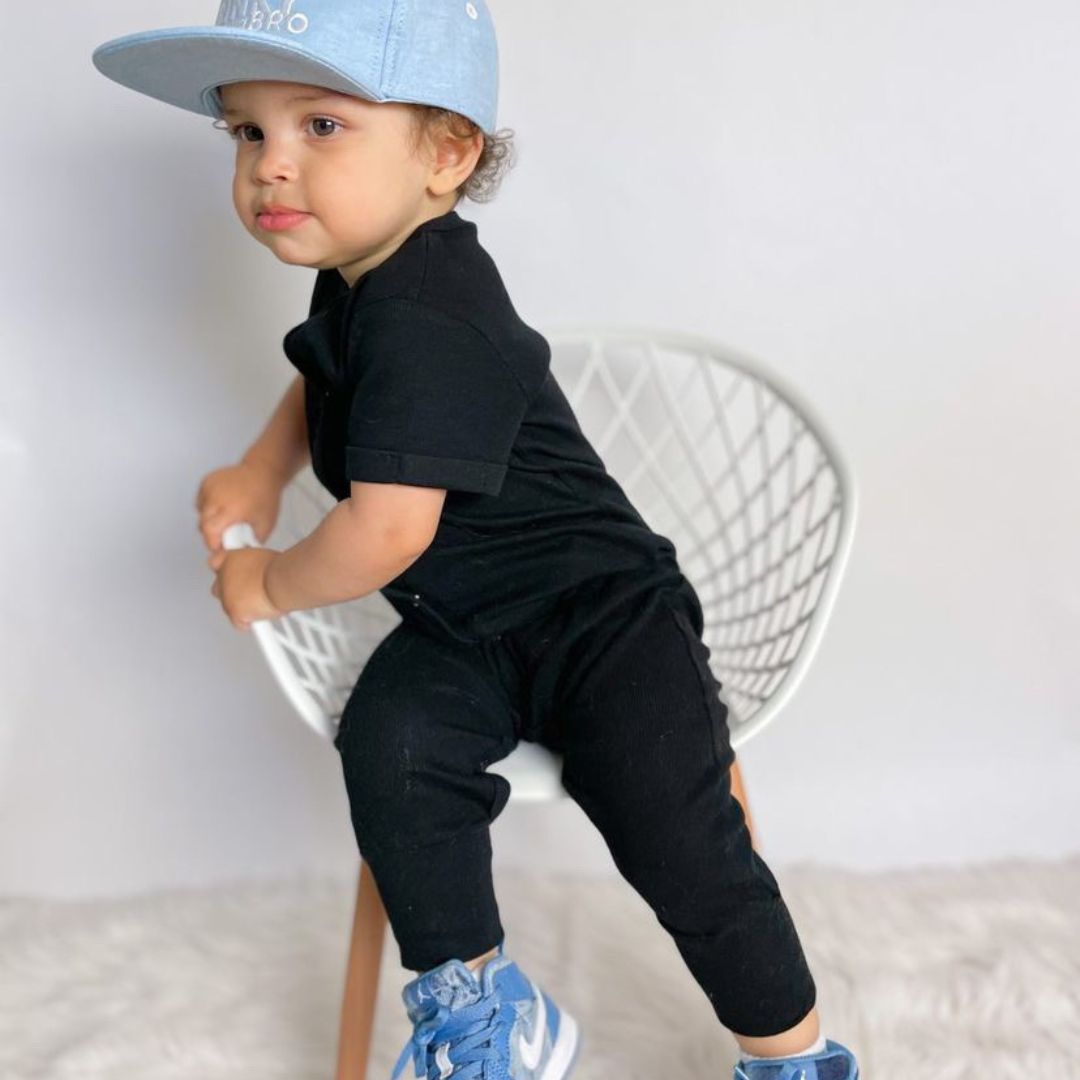 Toddler boy in a chair wearing the black ribbed romper and matching hat and kicks in baby blue.