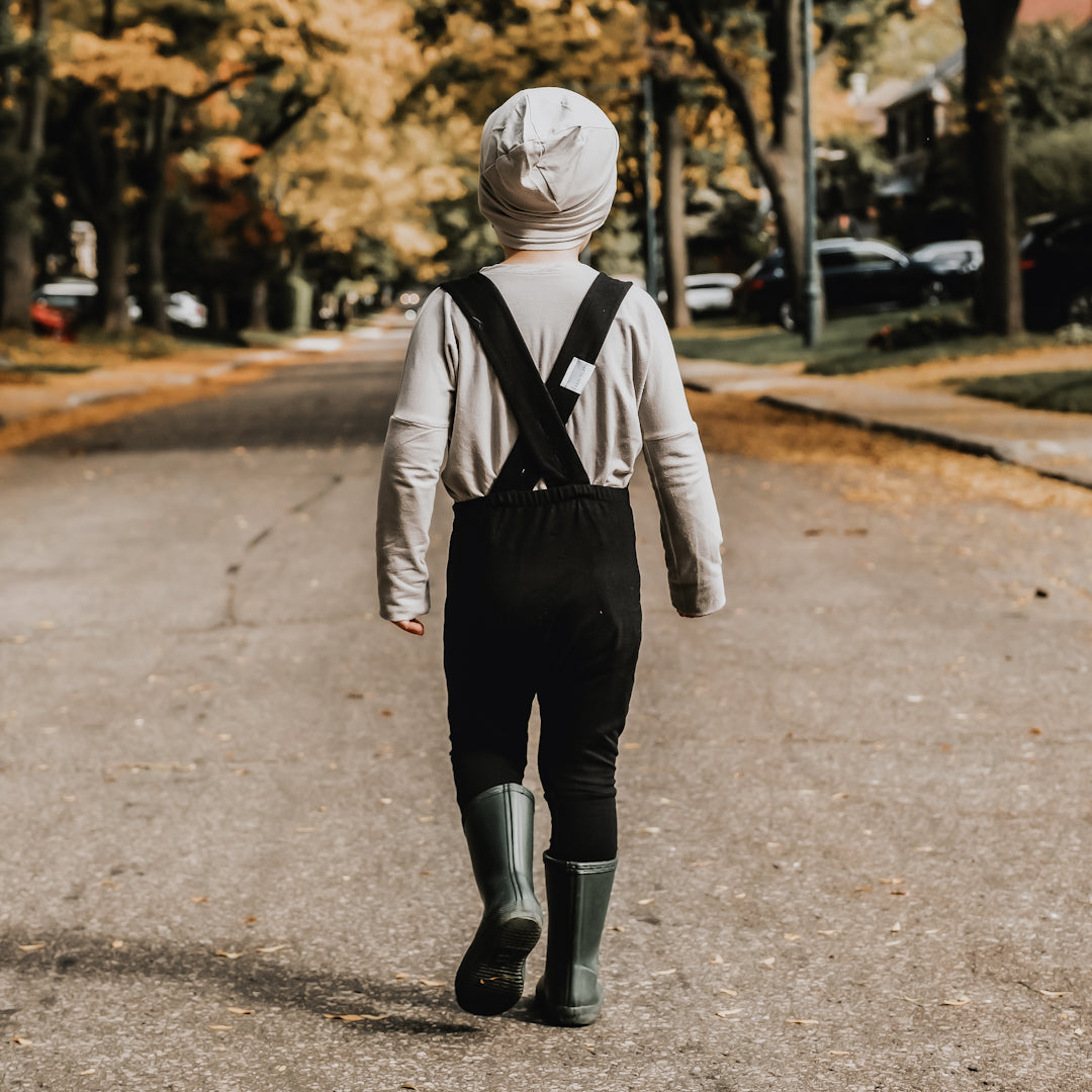 little boy walking away down a street covered with leaves wearing the grow with me overalls in black, black rain boots, and a toque and the grow with me shirt in taupe. He is showing the criss cross extendable straps. 