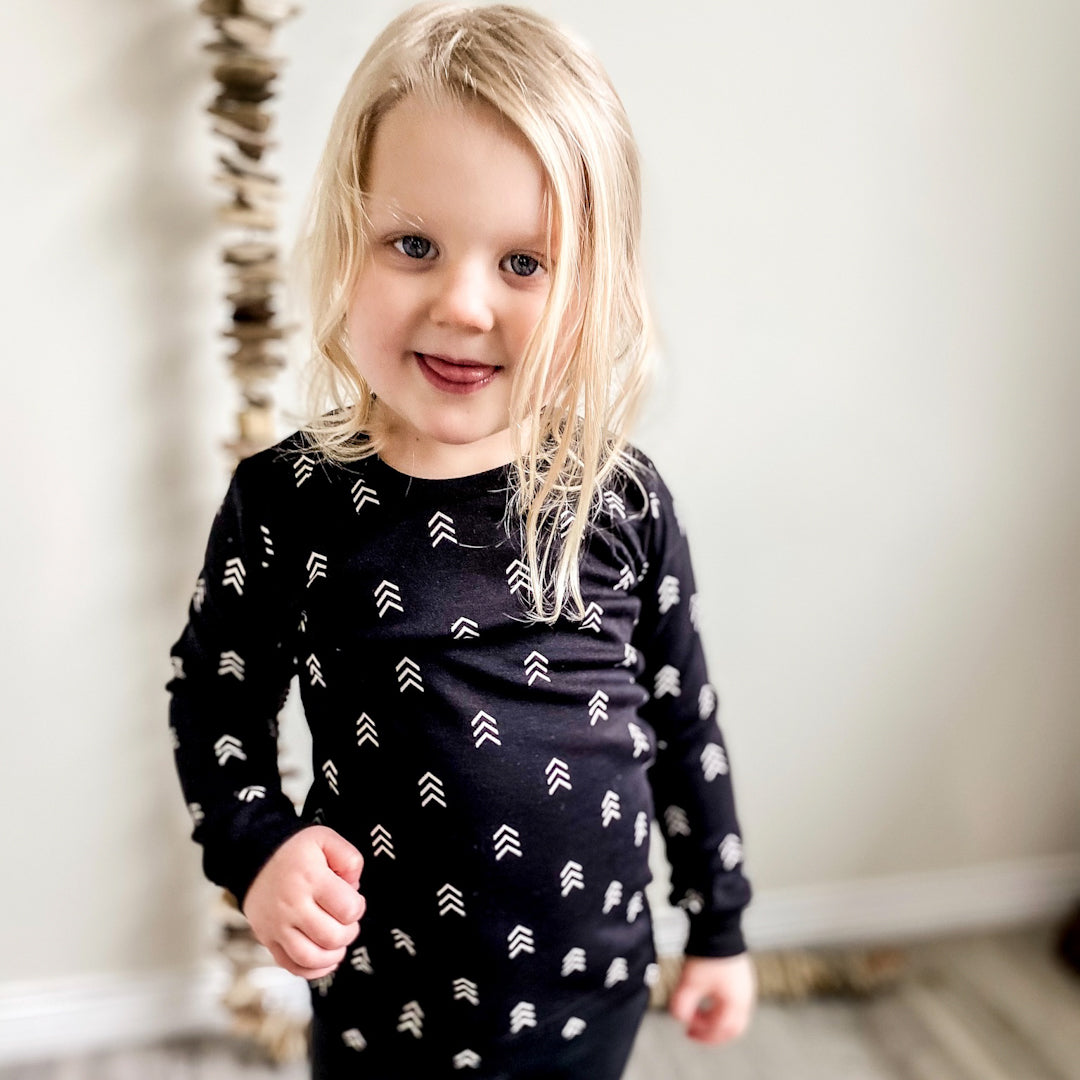 Blond girl facing the camera wearing a black long sleeved shirt with triple arrow pattern in white printed all over the shirt.
