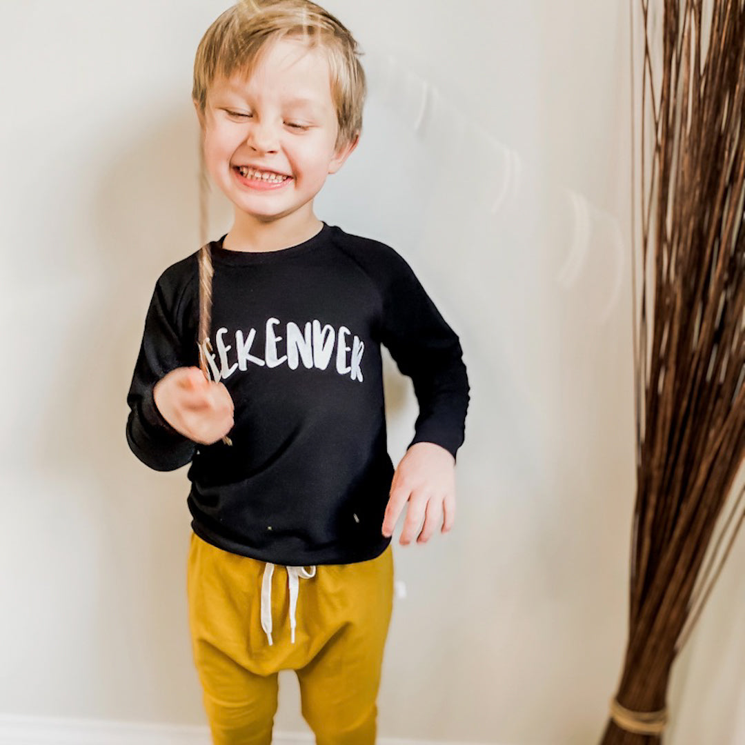 boy smiling holding a stick wearing the harem pants in golden yellow with the weekender raglan pullover in black