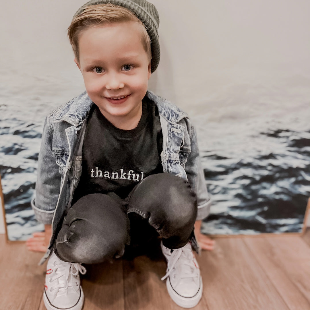 Blonde boy wearing the thankful shirt in heather charcoal black, black jeans, white shoes, and a green toque.