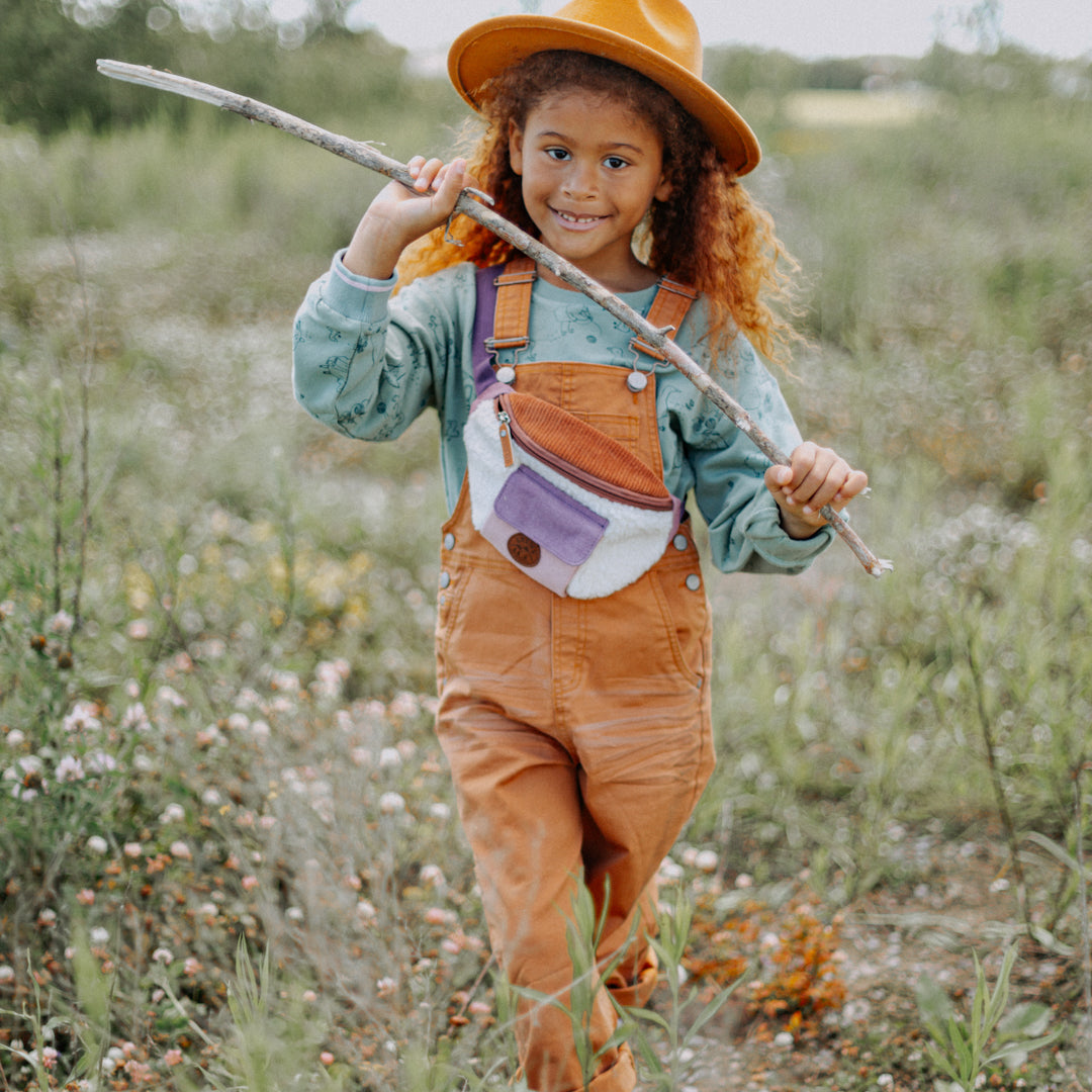 Little girl walking through a field holding a stick and wearing the Denim Overalls in Terracotta over a mint sweater.