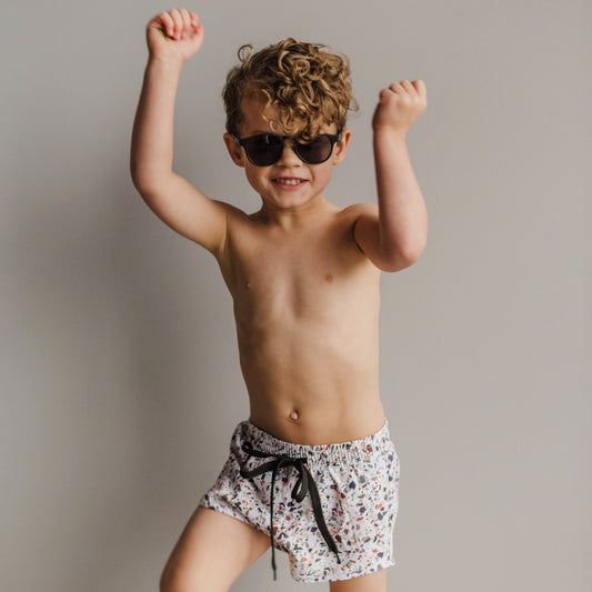 Little curly haired boy jumping in front of a light grey background wearing the Blake boardies, which have a multicoloured terracotta print