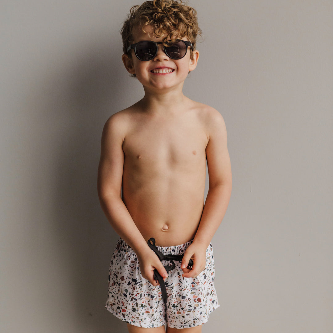 Little curly haired boy smiling in front of a light grey background wearing the Blake boardies, which have a multicoloured terracotta print