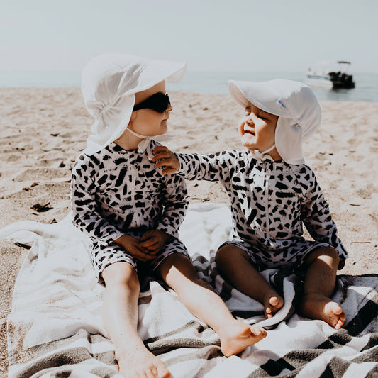 Two babies sitting on a beach blanket playing wearing the Iver One Piece Rash guard. It is a white suit with hand painted black splotches and a full length black zipper, full length sleeves, and knee length legs.