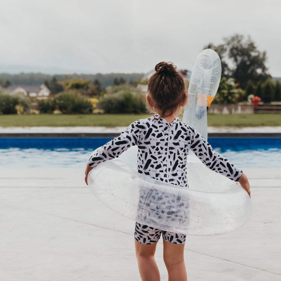 Little girl approaching the pool with a clear swan float. She is wearing the full sleeve length iver sunsuit with short length legs. It has a full zipper and is white with black paint splotches on it.
