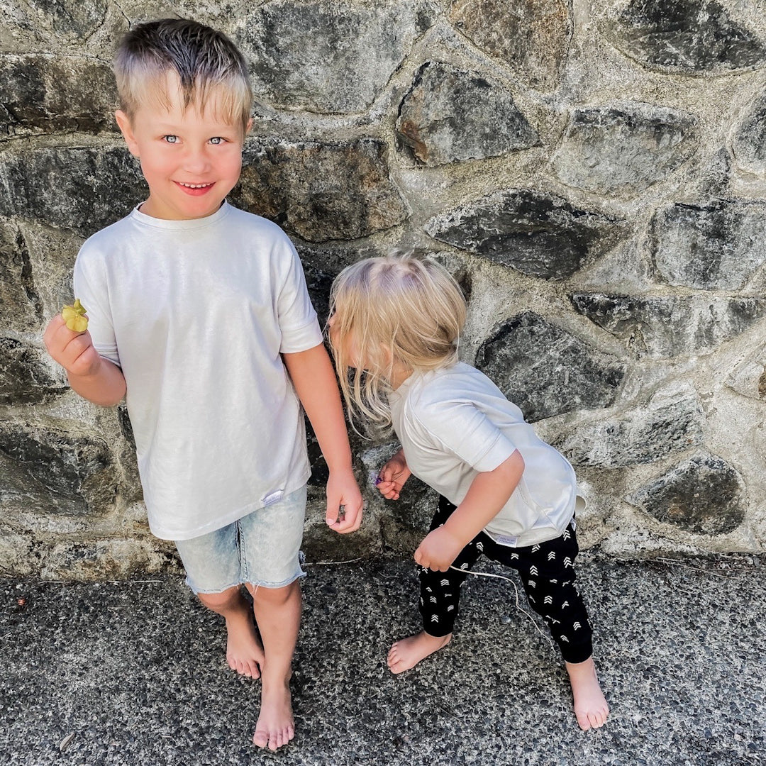 Little boy holding a flower with a little girl kissing his elbow. Both are wearing the grow with me shirt in taupe. His is paired with denim shorts and hers are paired with the harem pants with triple arrow print in black.