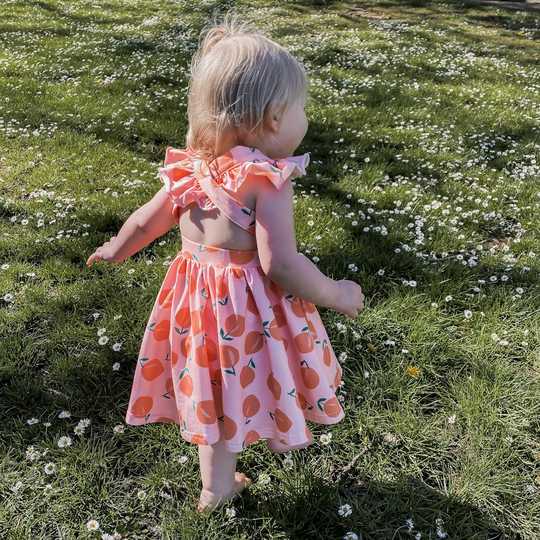 Baby girl walking away showing off the back of the Rosita dress in peachy.