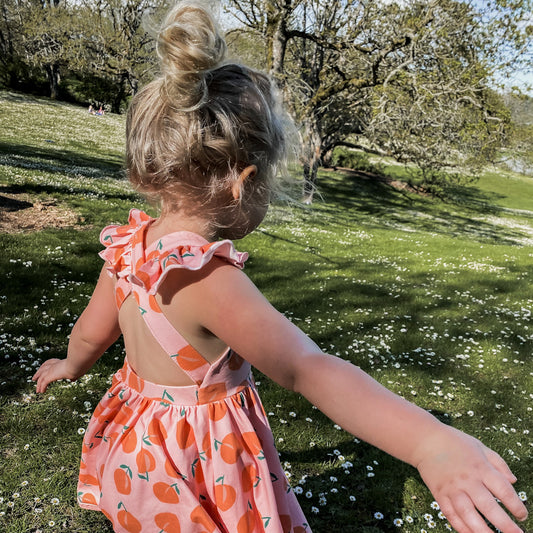 three year old girl wearing the Rosita dress in peachy. This showcases the ruffled criss crossed straps and open back.