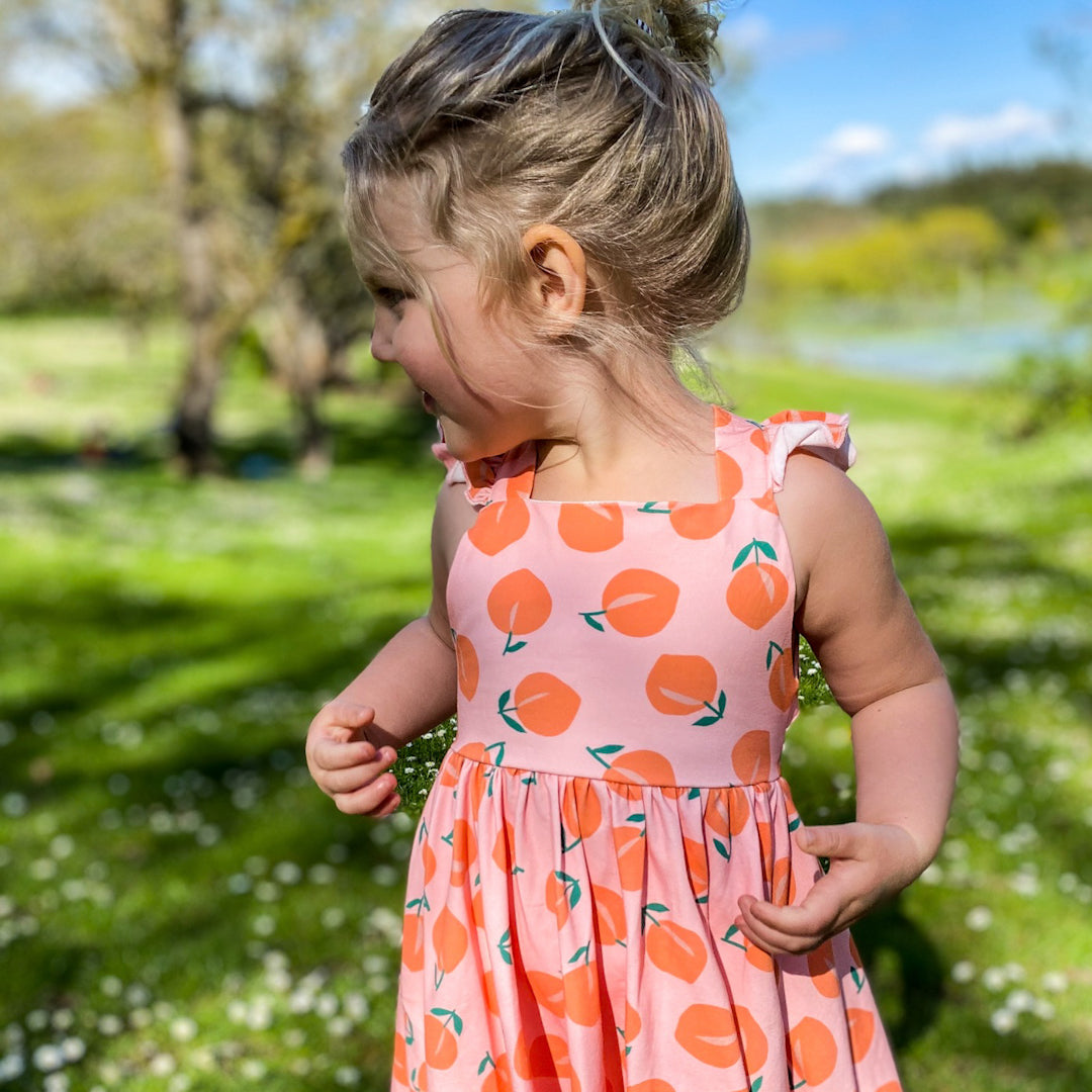 Blonde three year old wearing the Rosita dress in peachy. It is a peach coloured dress with a peach graphic all over.