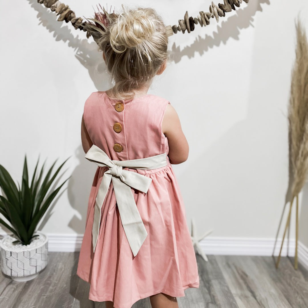 blonde girl showing the back of the pintuck dress in French pink with its wooden buttons and wheat coloured bow tied in the back.