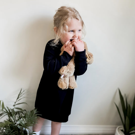 Blonde three year old girl wearing the raglan pullover sweater dress in black and holding a tan coloured bunny.  Edit alt text