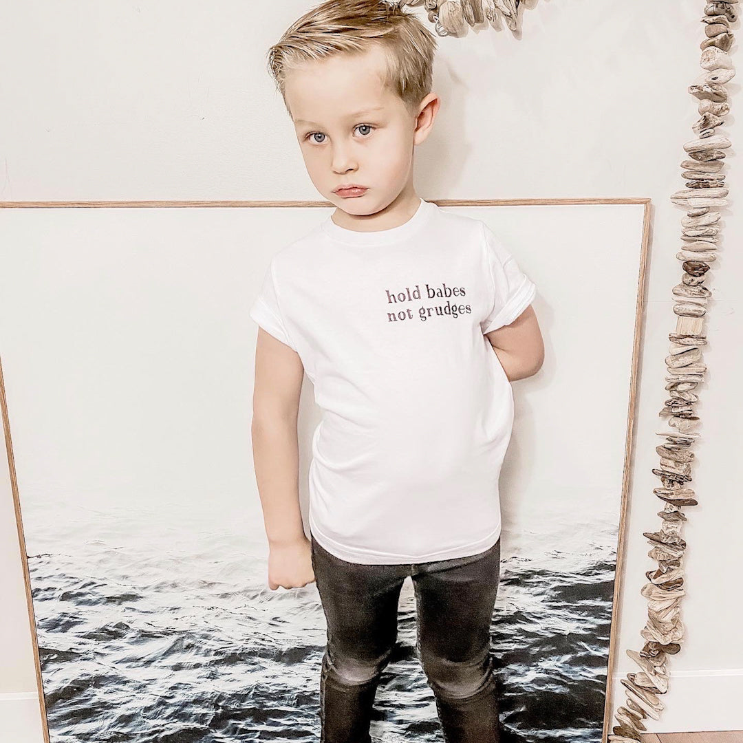 Toddler boy in a white t shirt with the words "Hold babes not grudges" written on the front. He is also wearing black distressed jeans, and white converse.