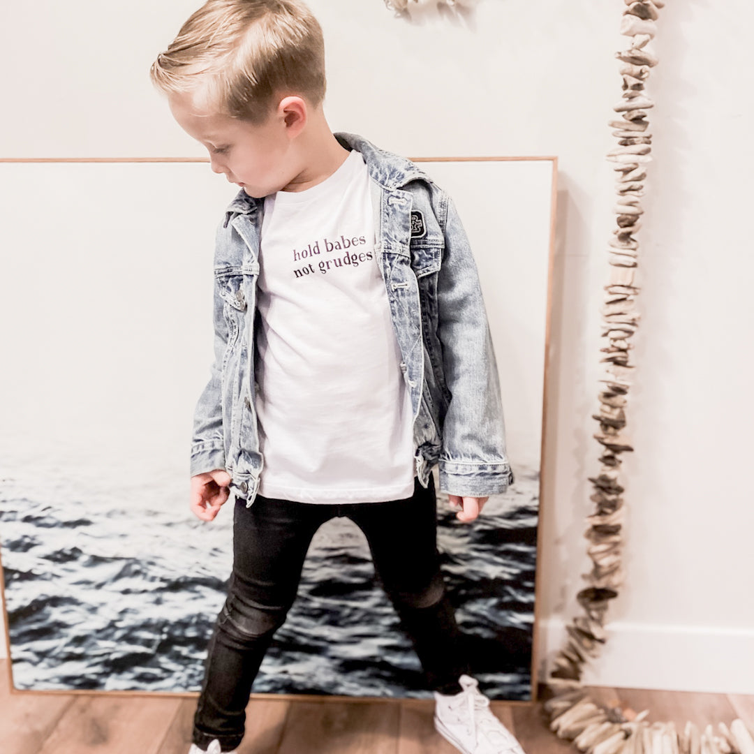 Toddler boy in a white t shirt with the words "Hold babes not grudges" written on the front. He is also wearing a blue denim jacket, black distressed jeans, and white converse.